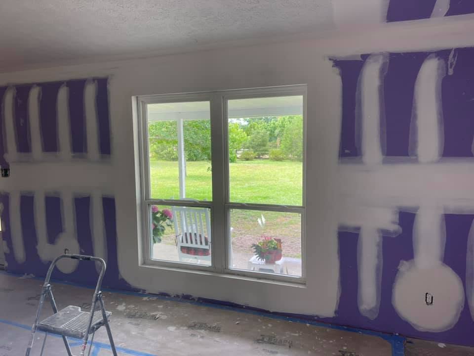 Interior room under renovation with purple drywall covered in white joint compound, featuring a large window looking out onto a green lawn and a step ladder on the floor.