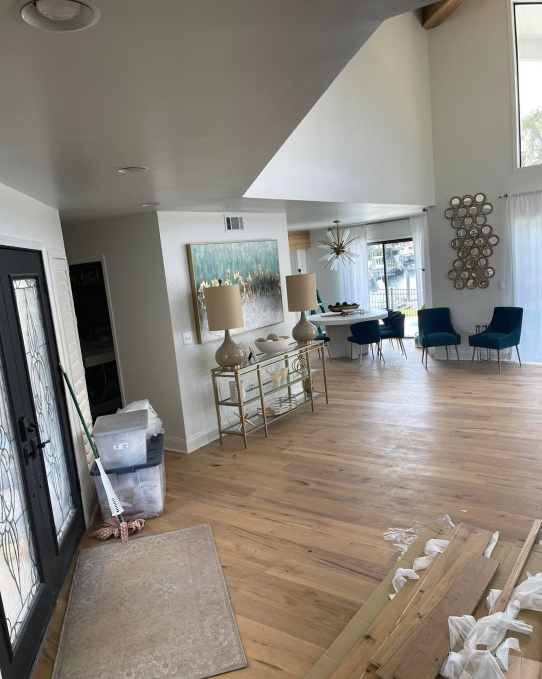 Modern living space with wooden flooring, blue chairs, a glass-console table with lamps, a round dining table, and large windows.