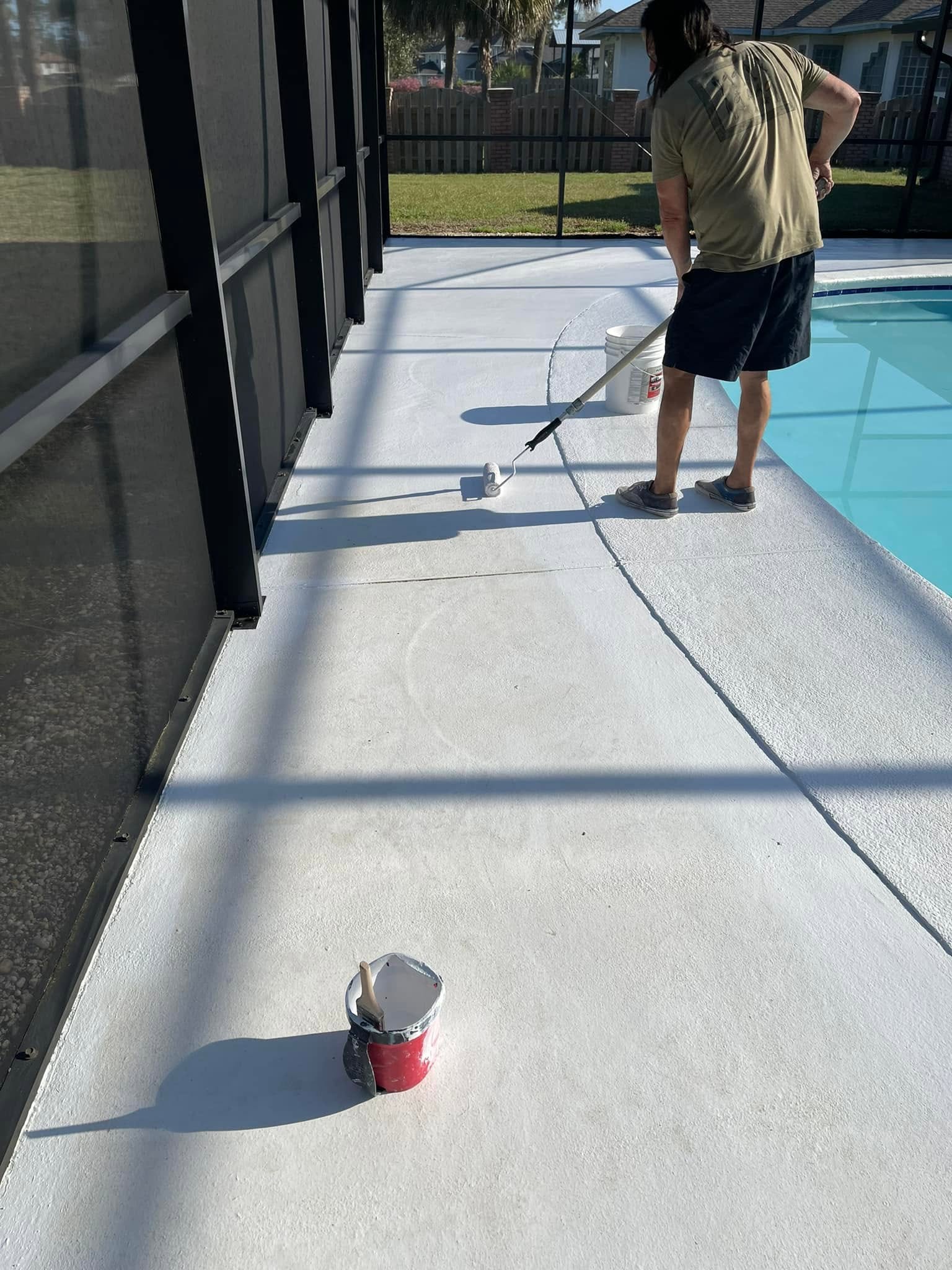Person using a paint roller to paint white concrete around a pool, with a paint bucket and brush visible on the ground nearby.