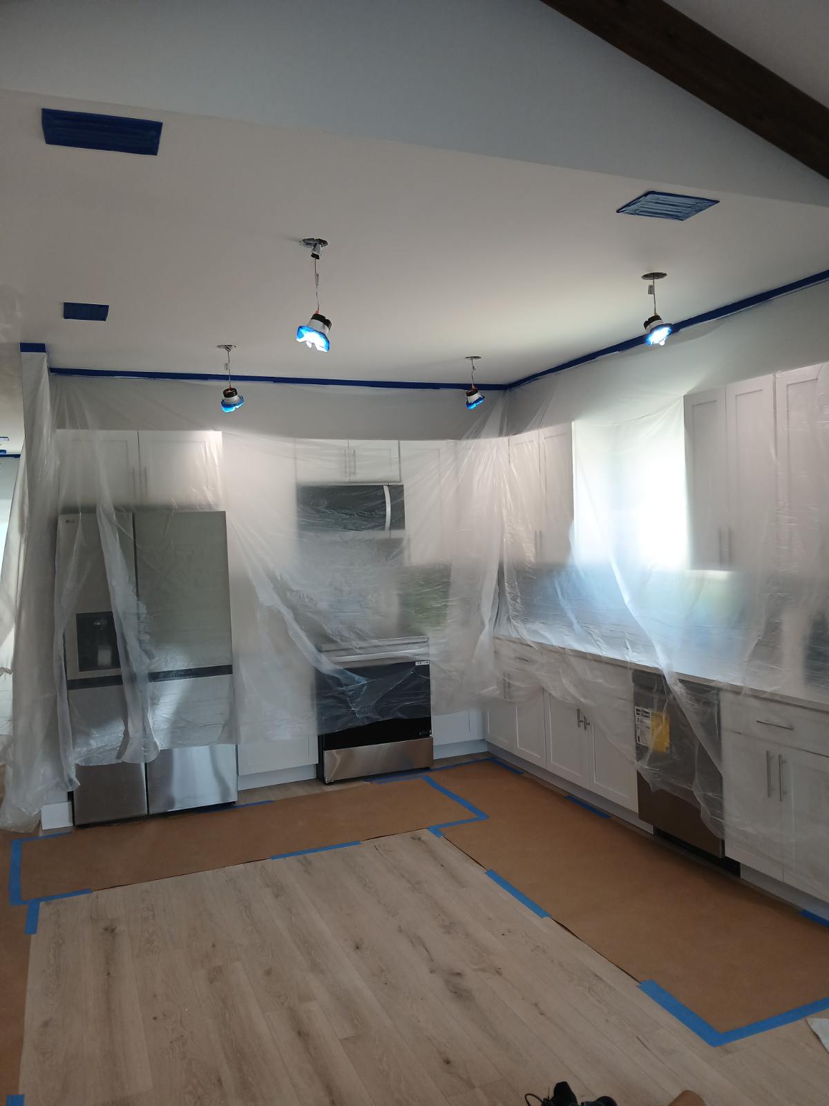 Kitchen under renovation, with cabinets and appliances wrapped in plastic, blue painter’s tape marking the floor, and bright work lights overhead.